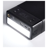 Ein schwarzes elektronisches Gerät mit Display, Knöpfen und mehreren Anschlüssen, darunter USB und DC Out, auf einer weißen Oberfläche.